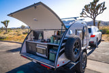 This RV rental teardrop camper, revealing its outdoor kitchen, is set up in a Joshua Tree landscape with an SUV.