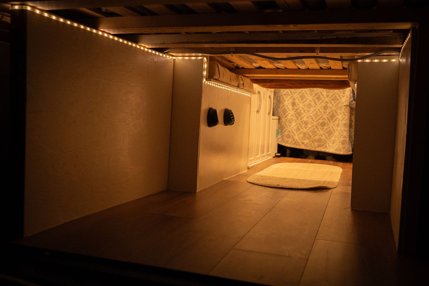 Functional RV rental under-bed storage area, illuminated by warm LED lights, with wooden floor and vents visible.