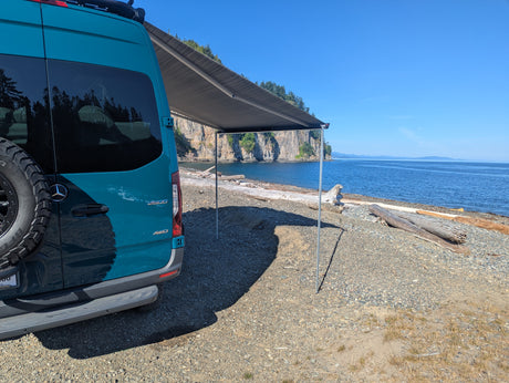 Teal RV rental with awning extended on a rocky beach by blue water & cliffs. Perfect for adventure!