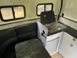 This RV rental interior shows a compact kitchen with stove/sink, black dinette seating, and bright windows.