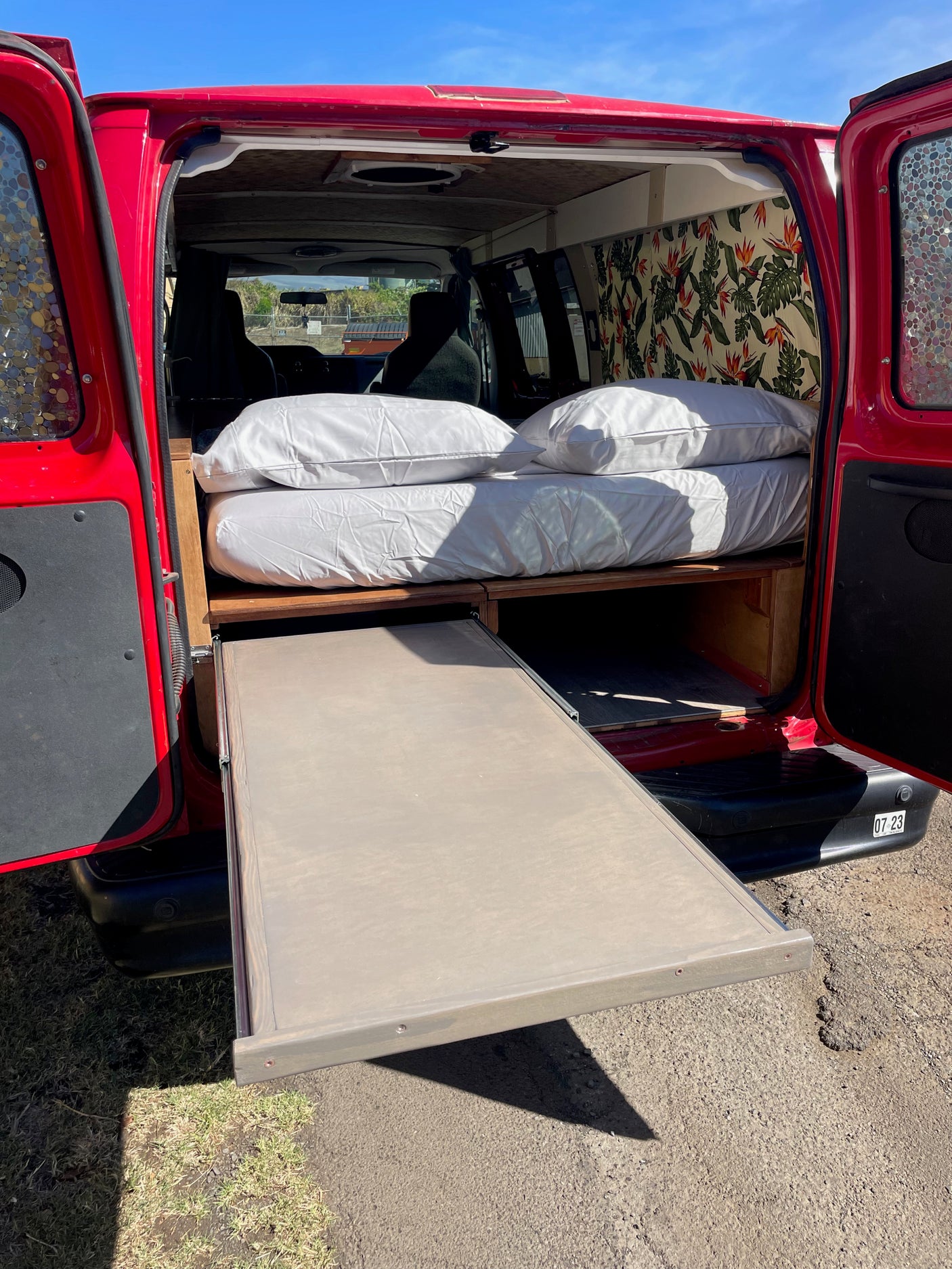 This rv rental camper features a cozy bed with pillows, patterned curtains, and a pull-out storage drawer.
