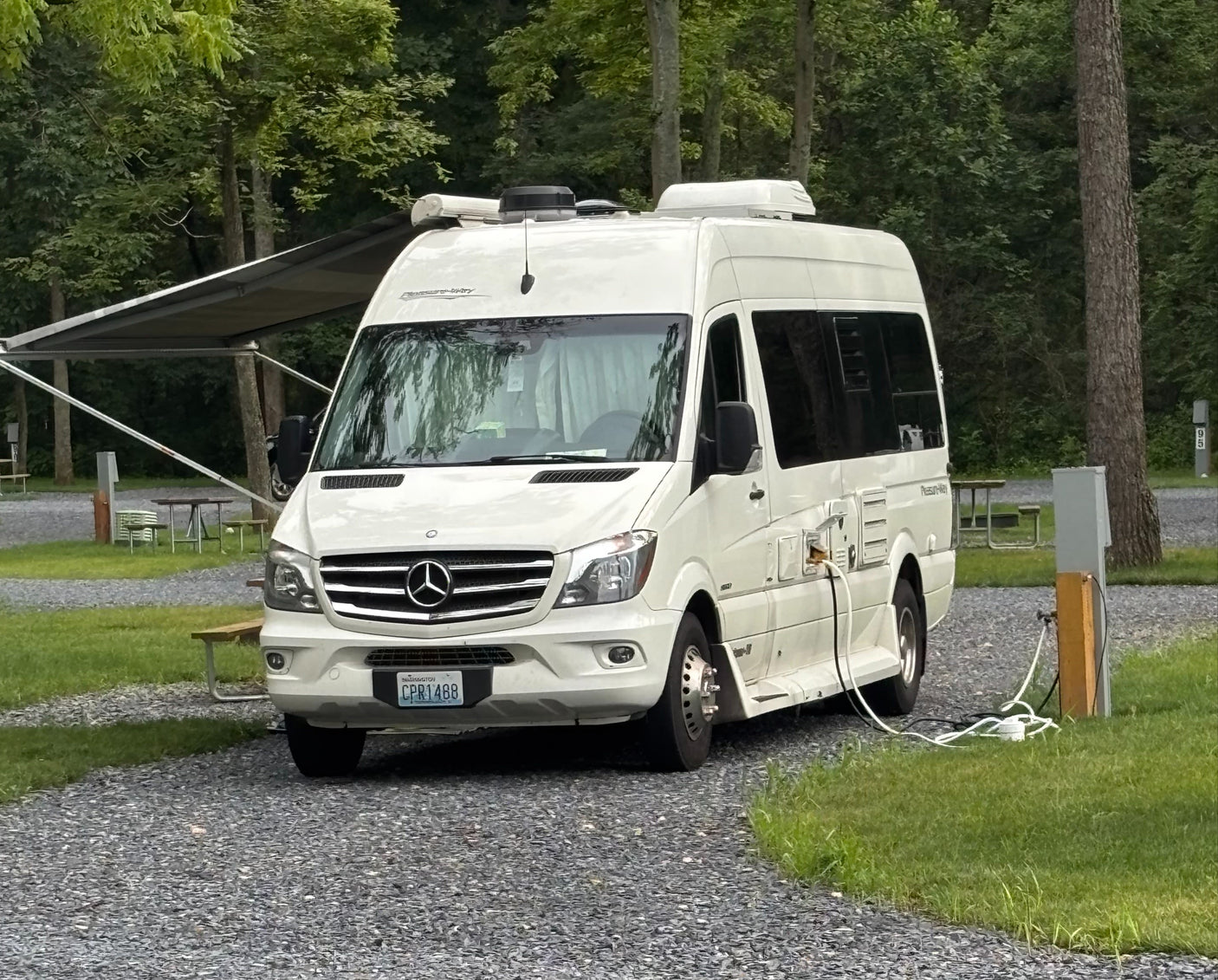A white Pleasure-Way RV rental is neatly parked and plugged in at a tranquil, wooded campsite, ready for adventure.