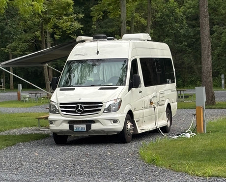 A white Pleasure-Way RV rental is neatly parked and plugged in at a tranquil, wooded campsite, ready for adventure.