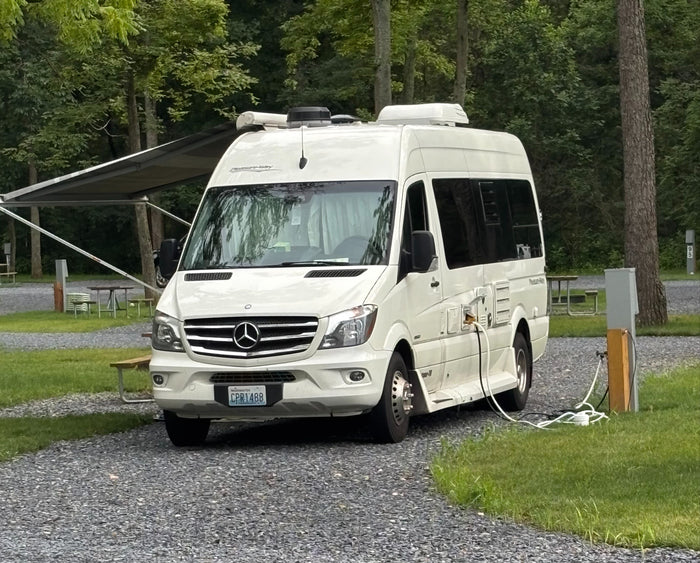 A white Pleasure-Way RV rental is neatly parked and plugged in at a tranquil, wooded campsite, ready for adventure.