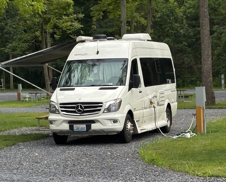 A white Pleasure-Way RV rental is neatly parked and plugged in at a tranquil, wooded campsite, ready for adventure.