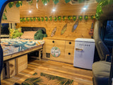 This RV rental interior features a cozy bed, mini-fridge, storage, and tropical decor with string lights & surfboards.