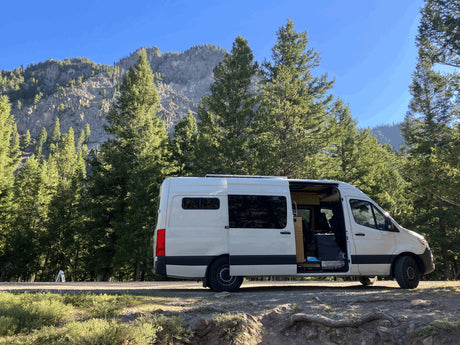 An RV rental camper van with its side door open, ready for adventure, parked among tall pines and rocky mountains.