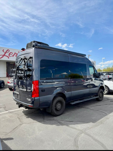 Dark gray Mercedes-Benz Sprinter RV, with a bike rack and awning, parked outdoors. Ideal for an rv rental adventure.