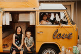 Family portrait with a vintage yellow Volkswagen rv rental. Two kids sitting in the open side, adults inside, 'love' sign.