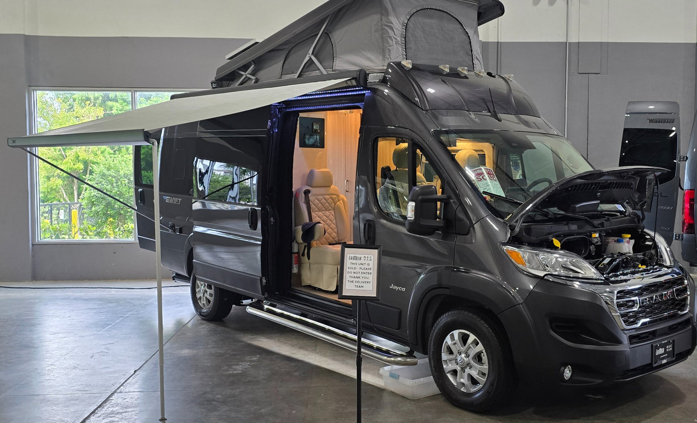 Dark gray Jayco camper van with pop-top and awning extended, interior illuminated and hood open, perfect for rv rental.