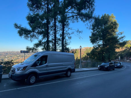 A grey Ford Transit van, suitable for an RV rental adventure, overlooks the Hollywood sign and Los Angeles cityscape.
