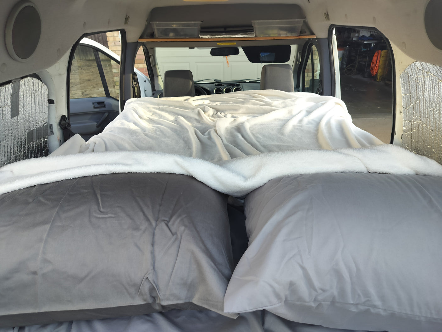 Inside an RV rental: a plush bed with grey pillows & white blanket, facing the front cabin. Ready for trips!