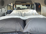 Inside an RV rental: a plush bed with grey pillows & white blanket, facing the front cabin. Ready for trips!