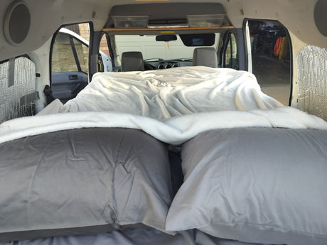 Inside an RV rental: a plush bed with grey pillows & white blanket, facing the front cabin. Ready for trips!