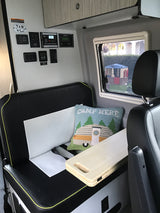 RV rental interior: comfy seating with a 'CAMP HERE' pillow, window view, and control panel for the camper van's systems.