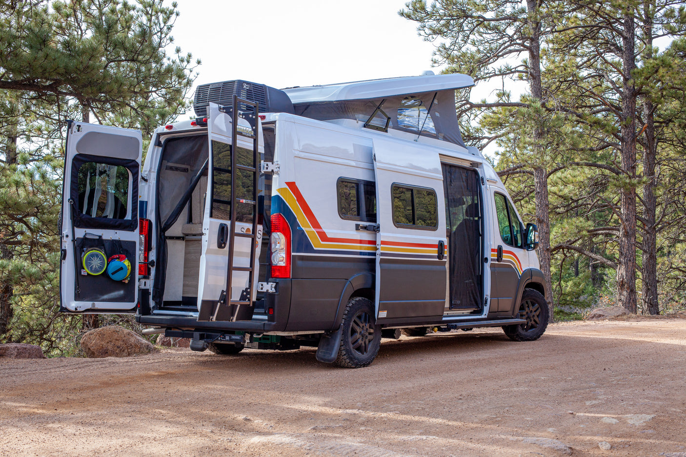 Adventure-ready RV rental camper van parked on a dirt road among pine trees with open doors & pop-top roof for outdoor living.