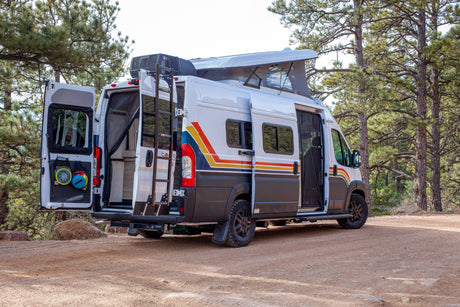 Adventure-ready RV rental camper van parked on a dirt road among pine trees with open doors & pop-top roof for outdoor living.