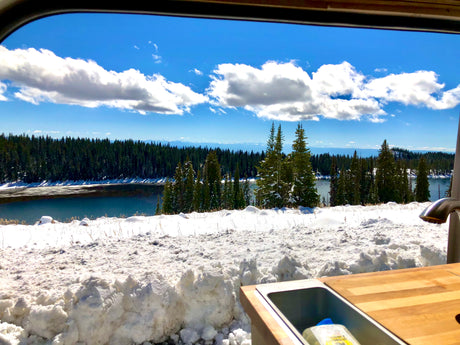 RV rental view: stunning snow-covered lake, forest, and blue sky. Cozy interior with sink, perfect winter adventure.