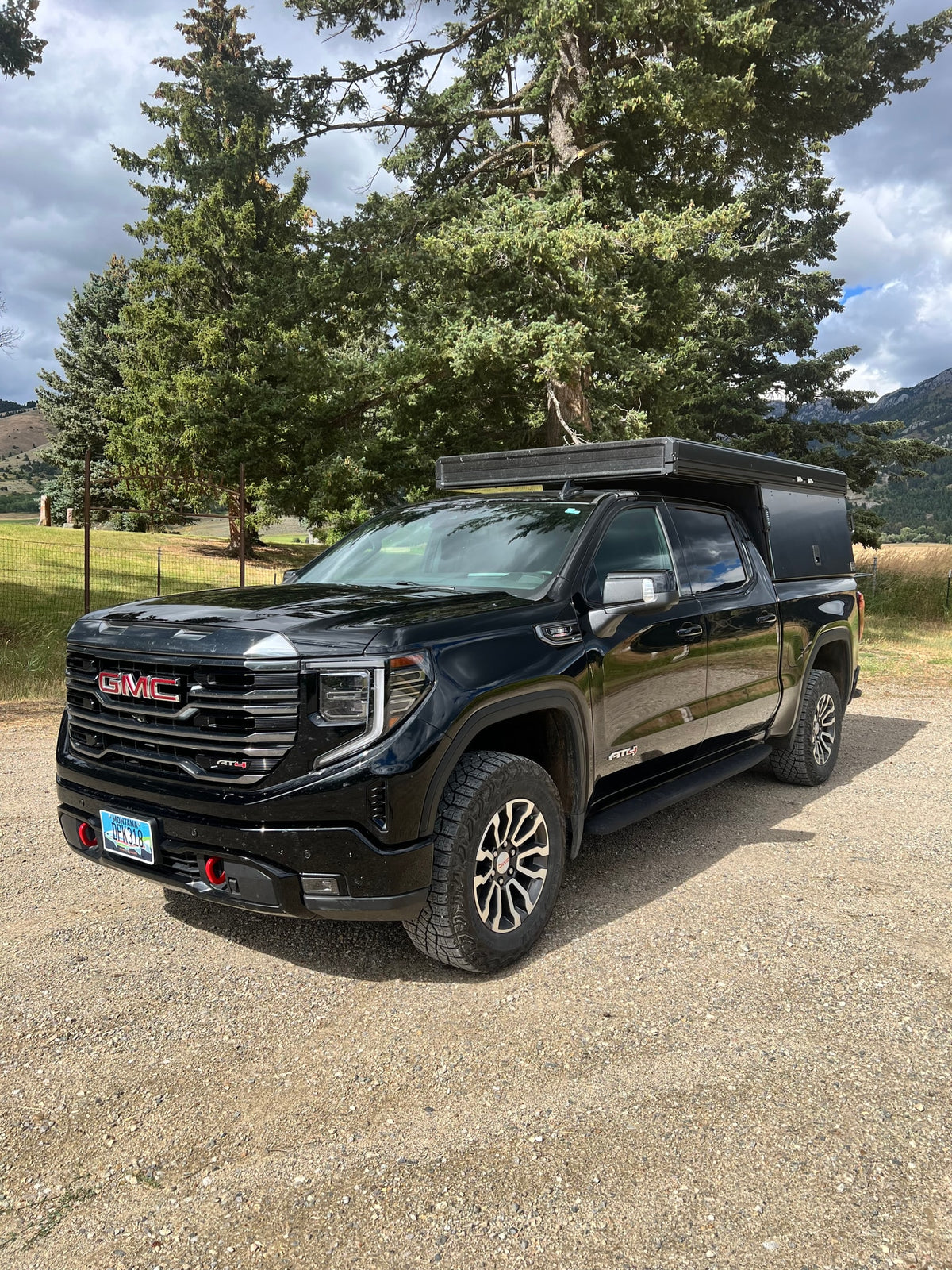 Black GMC Sierra AT4 pickup with a truck bed camper, parked on gravel with trees and mountains. Great for RV rental adventures.