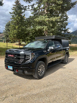 Black GMC Sierra AT4 pickup with a truck bed camper, parked on gravel with trees and mountains. Great for RV rental adventures.