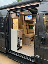 Step into this modern RV rental camper van! The open door reveals a sleek kitchen, cozy seating area, and TV inside.