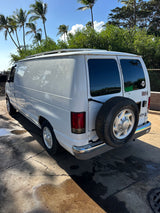 White RV rental van with a rear-mounted spare tire, parked on concrete under sunny palm trees.
