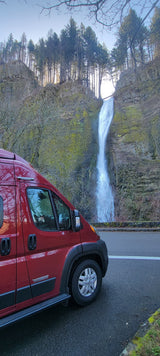 Red RV rental by a powerful waterfall cascading down a mossy cliff with tall trees. Perfect for a scenic outdoor adventure.