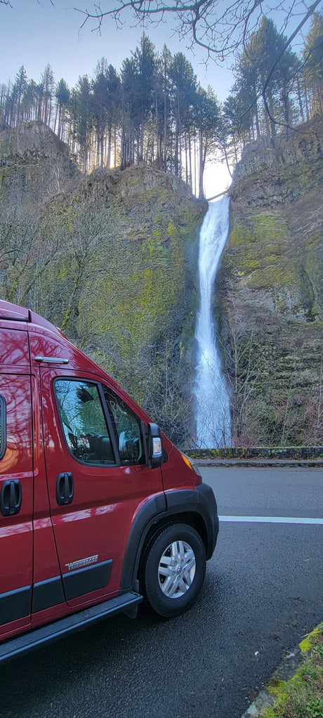 Red RV rental by a powerful waterfall cascading down a mossy cliff with tall trees. Perfect for a scenic outdoor adventure.