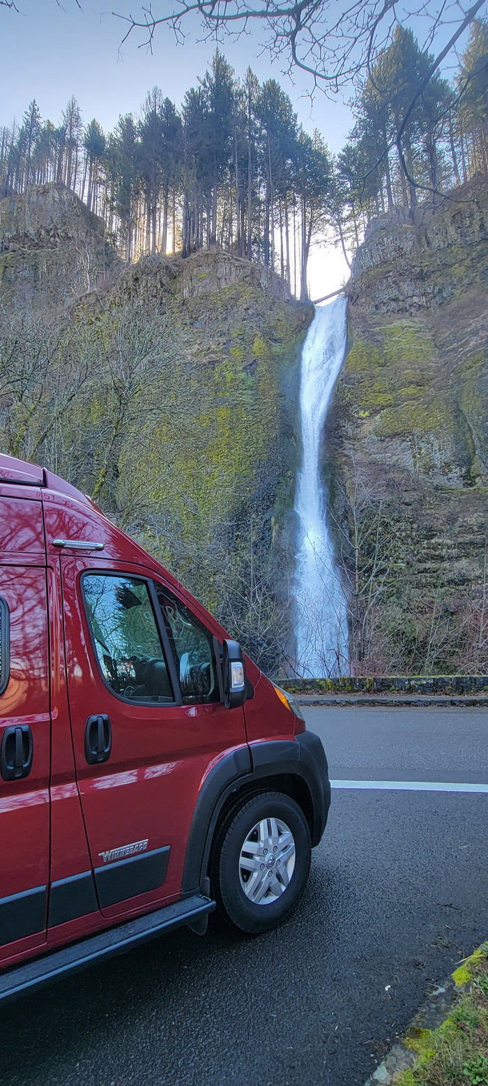 Red RV rental by a powerful waterfall cascading down a mossy cliff with tall trees. Perfect for a scenic outdoor adventure.