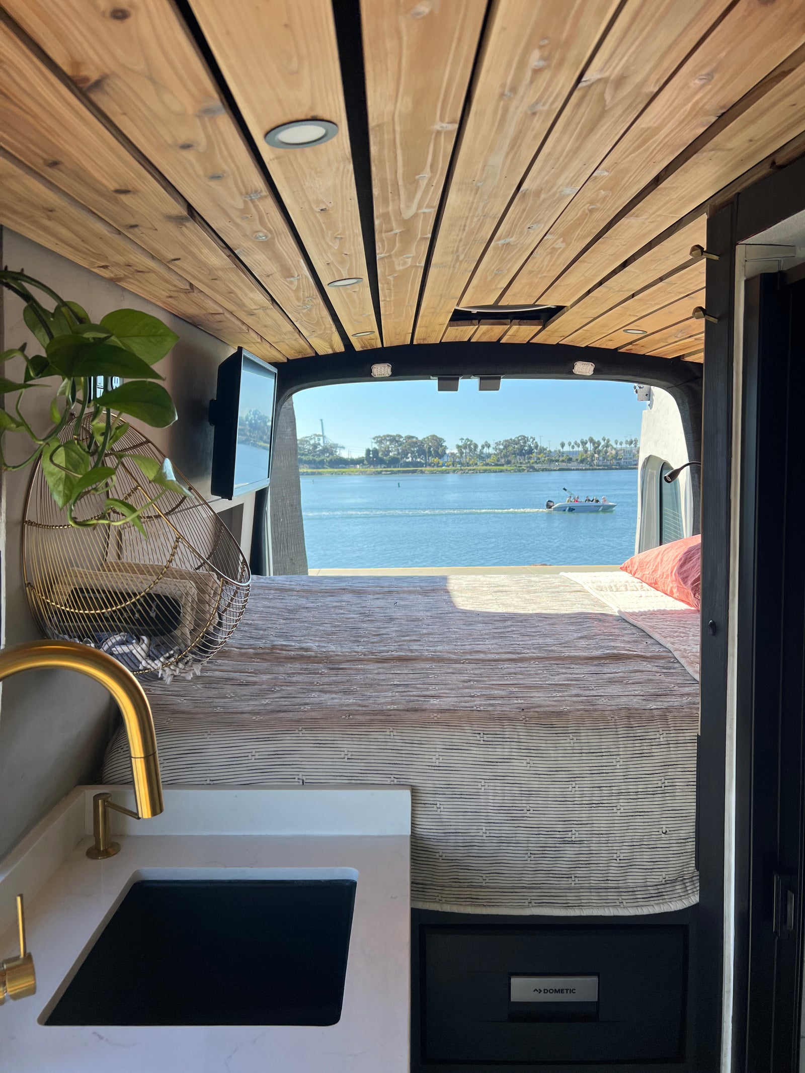 RV rental: Interior shot from the front showing a bed, kitchen counter with gold faucet, and a Dometic fridge, with a stunning water view from the rear.