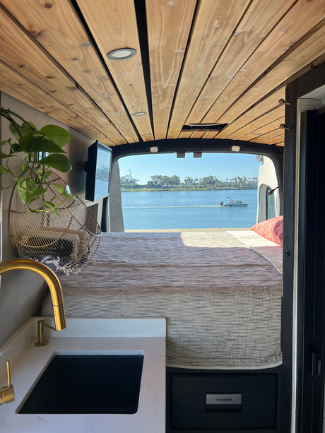 RV rental: Interior shot from the front showing a bed, kitchen counter with gold faucet, and a Dometic fridge, with a stunning water view from the rear.