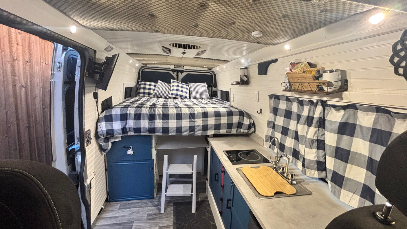 RV rental camper van interior: features a raised bed with plaid bedding, kitchenette with sink/cooktop, and wall-mounted TV.