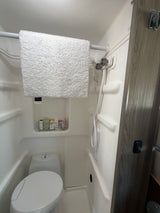 Compact RV rental bathroom featuring a shower, toilet, and toiletries. A white towel hangs on a rod above the toilet.