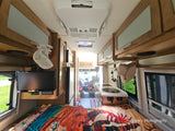 Interior of an RV rental, looking forward from the bed to the kitchenette, overhead cabinets, and driver's cockpit.