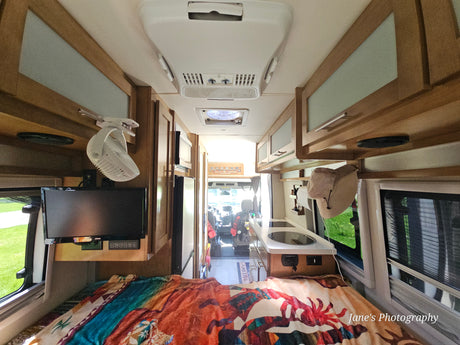 Interior of an RV rental, looking forward from the bed to the kitchenette, overhead cabinets, and driver's cockpit.