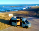 Aerial view of a modern RV rental parked on a sandy beach by the ocean with a rock jetty. Its open door shows interior.
