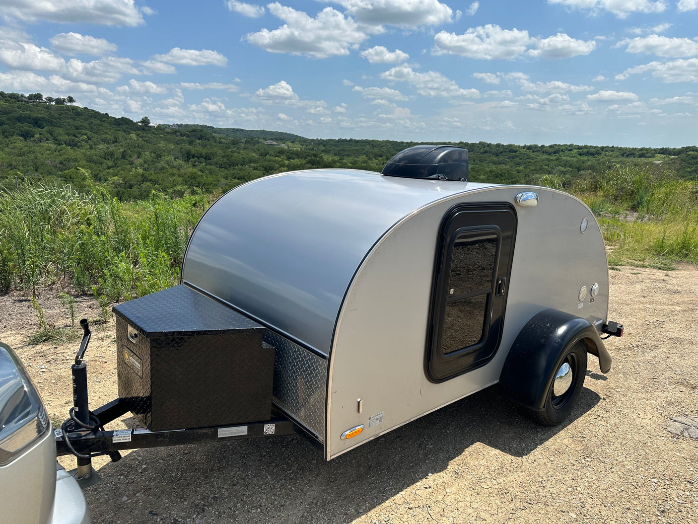 Compact RV rental: A silver teardrop trailer on a dirt path with green hills & a bright blue sky overhead.