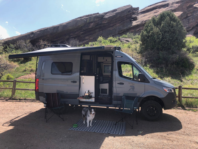 Grey camper van RV rental parked by scenic red rocks. Features open side with awning, chairs, dog & outdoor kitchen. — photo 9