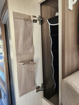 Inside an RV rental: an open overhead cabinet reveals a black foldable organizer. The door has a beige mesh pocket system, above a window.