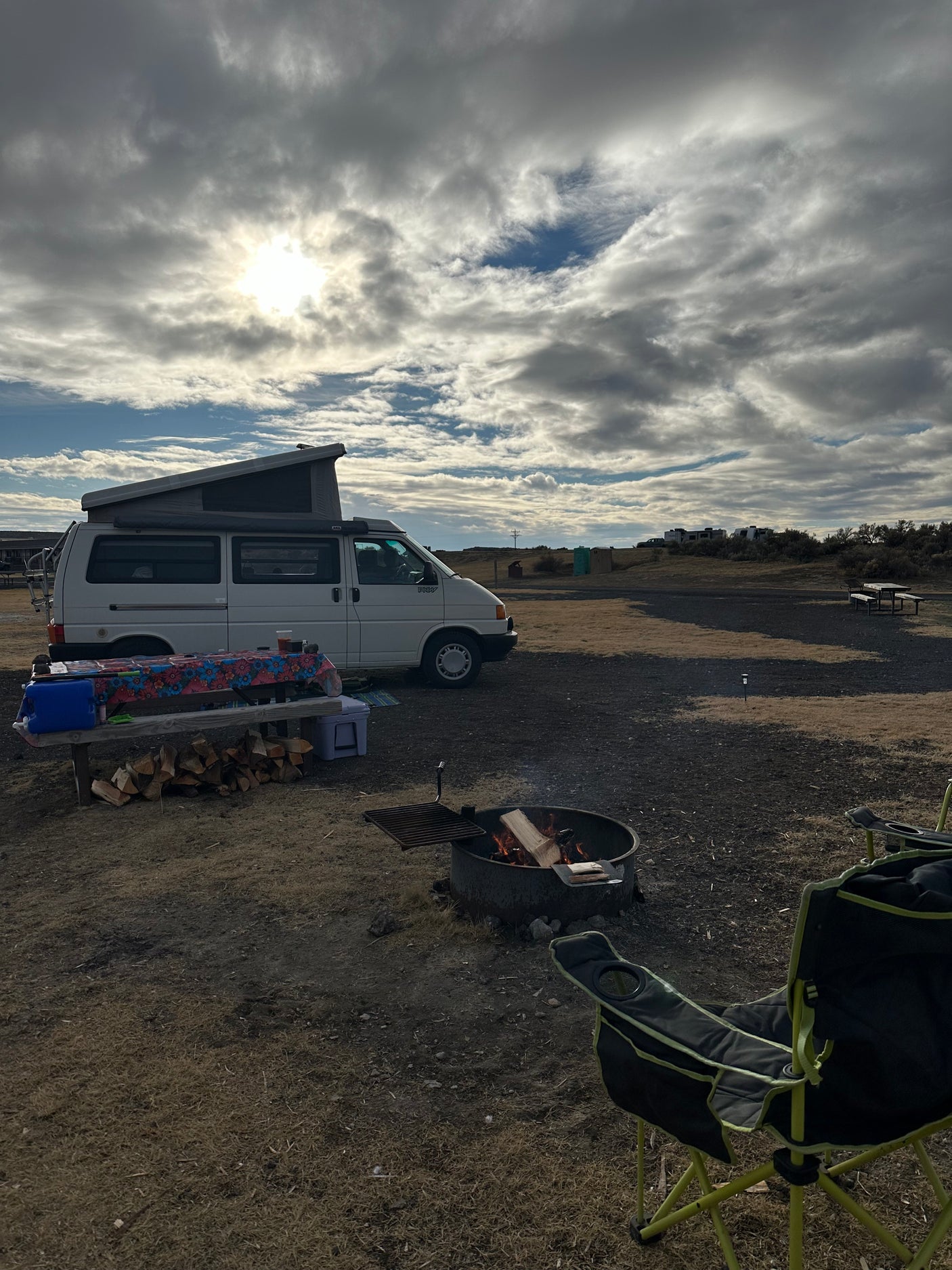 Camper van RV rental at a campsite with a roaring campfire, picnic table, and chair under a dramatic cloudy sky.