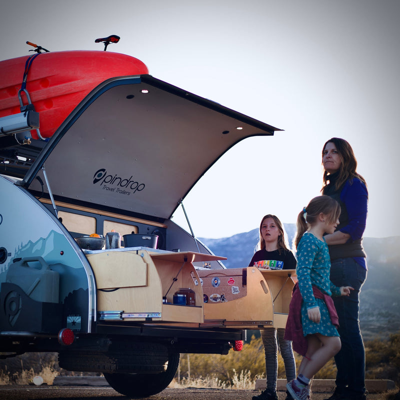  parked in a vast desert. An ideal RV rental for off-road camping.|An RV rental alternative: a family with their Pindrop teardrop travel trailer and open kitchenette for camping.|Enjoying an RV rental experience: Family cooks outdoors from their Pindrop teardrop camper — photo 2
