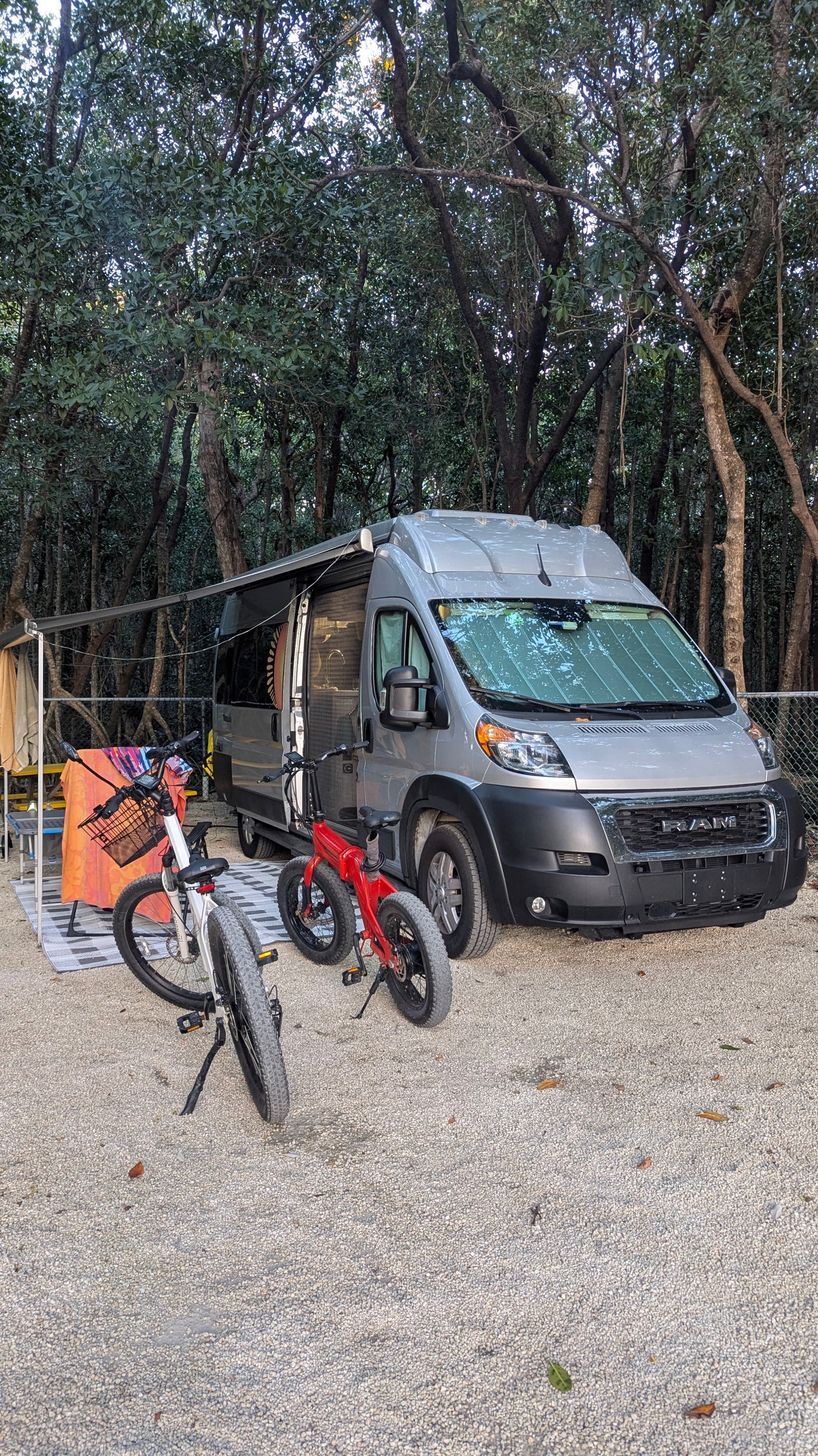 This silver RV rental is set up at a shady, treed campsite on gravel, with an awning extended and two bicycles parked nearby.