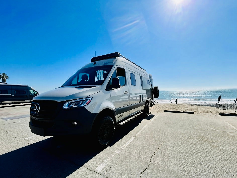 Adventure awaits! White Mercedes-Benz Sprinter rv rental at a sunny beach with the ocean, waves, and people.