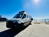 Adventure awaits! White Mercedes-Benz Sprinter rv rental at a sunny beach with the ocean, waves, and people.