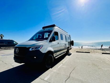 Adventure awaits! White Mercedes-Benz Sprinter rv rental at a sunny beach with the ocean, waves, and people.