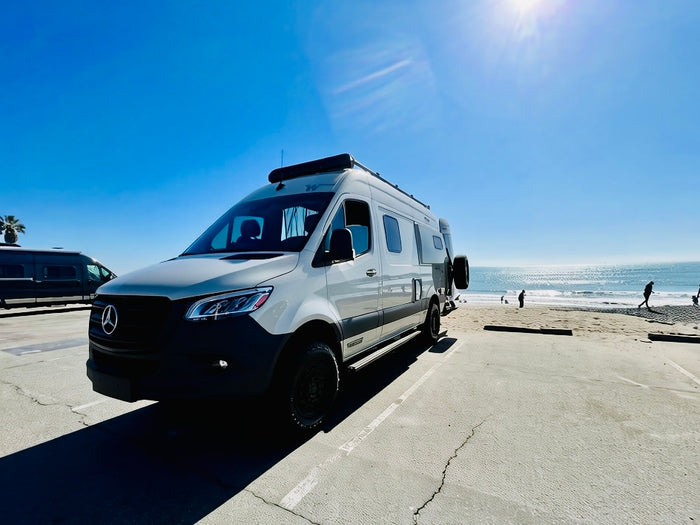 Adventure awaits! White Mercedes-Benz Sprinter rv rental at a sunny beach with the ocean, waves, and people.