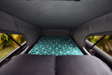 Cozy RV rental pop-top bed setup with a teal geometric blanket and two dark pillows. Lush green foliage visible outside.
