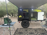 Adventure-ready RV rental setup: black Outbound T-Rex camper trailer, awning, camp kitchen, and chairs on gravel.