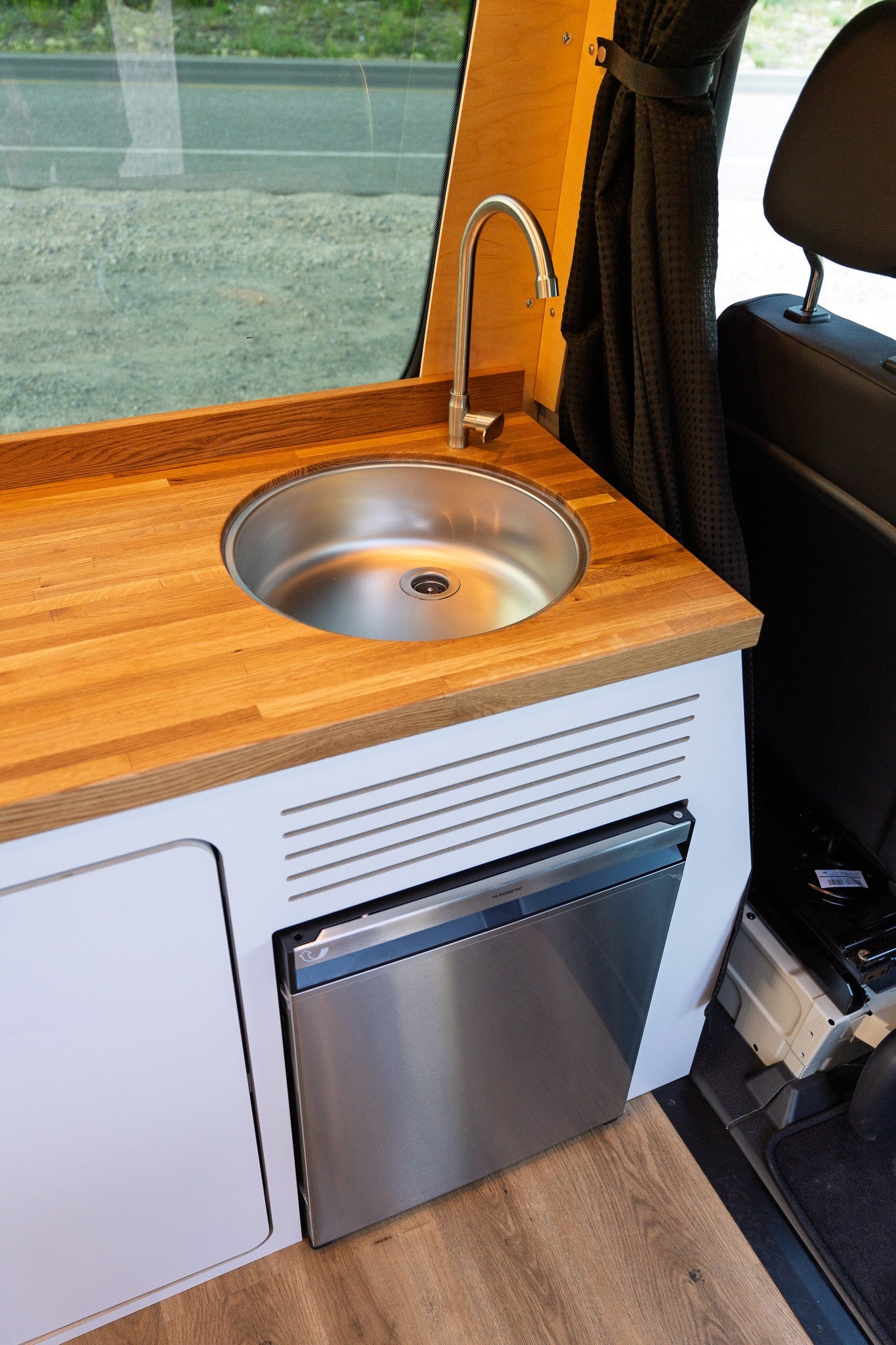 RV rental kitchen: wood counter, stainless sink, faucet, & compact Dometic fridge in a modern camper van.