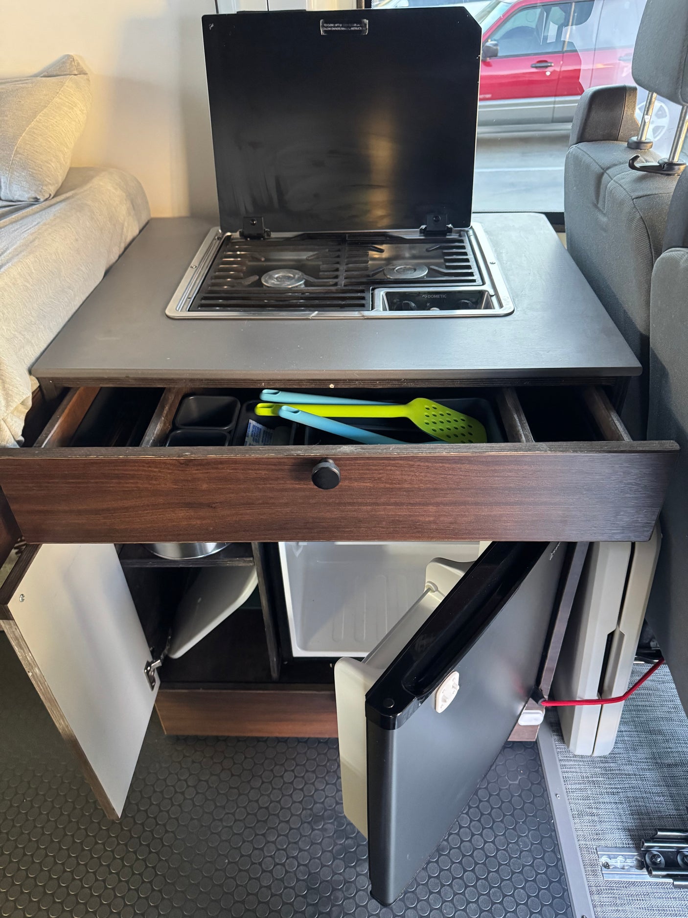 Compact RV rental kitchen with Dometic stove, open drawer with utensils, and mini-fridge. Ideal for camper cooking.